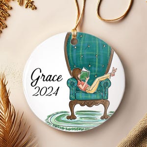 May include: A round ceramic ornament with a watercolor illustration of a person reading in a teal armchair. The ornament has the text "Grace 2024" in black script. A gold string is attached for hanging.