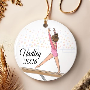 May include: A round white ceramic ornament featuring a printed illustration of a gymnast in a pink leotard performing on a balance beam. The text "Hadley" and "2026" is printed in black script and serif font. A gold string is attached for hanging.