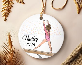 May include: A round white ceramic ornament featuring a printed illustration of a gymnast in a pink leotard performing on a balance beam. The text "Hadley" and "2026" is printed in black script and serif font. A gold string is attached for hanging.