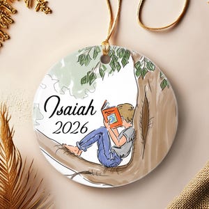 May include: A round white ceramic ornament featuring a printed illustration of a person sitting in a tree reading an orange book. The text Isaiah and 2026 are printed in black script and sans-serif font on the left side of the ornament.