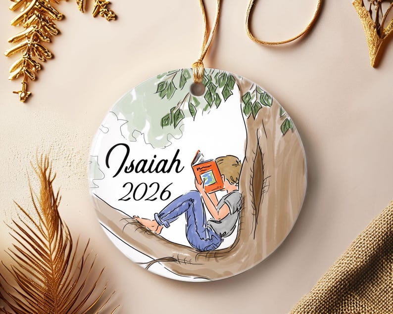 May include: A round white ceramic ornament featuring a printed illustration of a person sitting in a tree reading an orange book. The text Isaiah and 2026 are printed in black script and sans-serif font on the left side of the ornament.