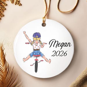 May include: A round white ceramic ornament featuring a colorful illustration of a person with blonde hair in pigtails and a purple cap riding a bicycle. The text Megan and 2026 is printed in black script and sans-serif font on the right side.