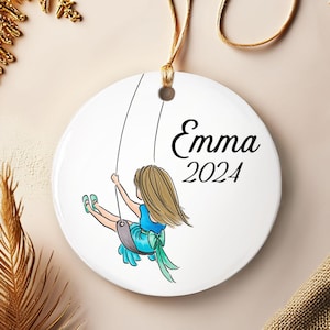 May include: White ceramic ornament featuring a girl on a swing, with the name "Emma" and the year "2024" in black script. The girl is wearing a blue dress and has blonde hair. The ornament has a gold string for hanging.