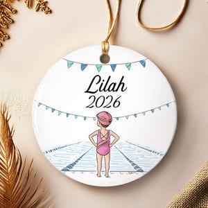 May include: A round white ceramic ornament featuring a printed illustration of a swimmer in a pink swimsuit and cap standing at the edge of a pool. The text Lilah and 2026 is printed in black above the swimmer, with blue bunting flags draped above.