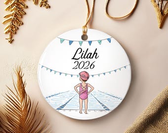 May include: A round white ceramic ornament featuring a printed illustration of a swimmer in a pink swimsuit and cap standing at the edge of a pool. The text Lilah and 2026 is printed in black above the swimmer, with blue bunting flags draped above.