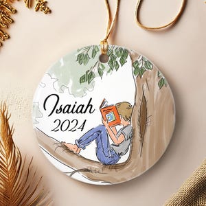 May include: Round ceramic ornament featuring a child reading a book while sitting in a tree. The ornament has the name "Isaiah" and the year "2024" printed on it. The ornament is suspended by a gold cord.