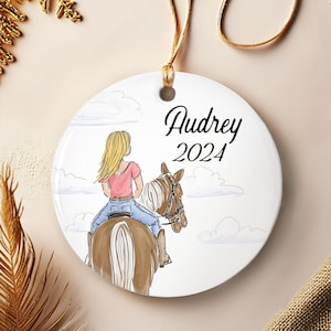 May include: A round, white ceramic ornament with a watercolor illustration of a person riding a brown horse. The ornament has the name "Audrey" and the year "2024" written in black script. A gold string is attached for hanging.