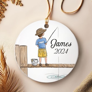 May include: A round ceramic ornament featuring a cartoon illustration of a person fishing off a wooden dock. The person is wearing a blue shirt, yellow shorts, and a brown hat. The ornament has the name "James" and the year "2024" written on it.