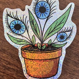 May include: A holographic sticker featuring a potted plant with three blue eyes. The plant has green leaves and is in an orange glitter pot. The eyes have black pupils and long eyelashes. The sticker has a silver border.