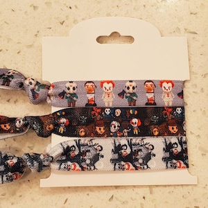 Puede incluir: Tres gomas para el pelo de colores con temática de películas de terror. Las gomas para el pelo presentan varios personajes de películas de terror, incluidos Freddy Krueger, Jason Voorhees y Pennywise el payaso bailarín.