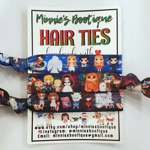 Könnte beinhalten: Drei bunte Haargummis mit verschiedenen Comicfiguren darauf gedruckt. Die Haargummis werden auf einer weißen Karte mit dem Text "Minnie's Boutique Hair Ties Handmade with ♥" und den Kontaktinformationen des Shops präsentiert.