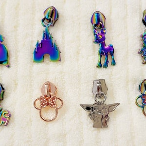May include: Assortment of colorful zipper pulls featuring various cartoon characters, including a deer, a castle, a snowman, a mushroom, a baby, and an alien. The zipper pulls are made of metal and have a rainbow iridescent finish.