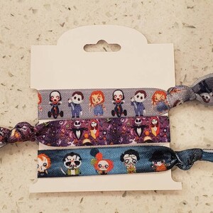 May include: Three colorful hair ties with different cartoon characters printed on them. The hair ties are on a white card with a hole at the top for hanging.