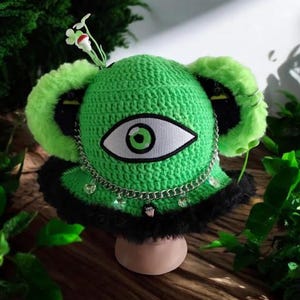 May include: A bright green crocheted bucket hat featuring a large central eye patch, fuzzy lime green ear-like accents, and a black faux fur brim. A silver-toned metal chain drapes across the front, adorned with small clear charms and a tiny black lip charm.