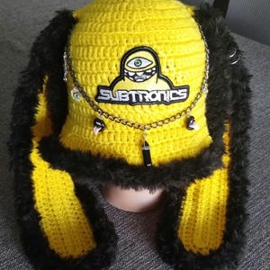 May include: A yellow crochet hat with black faux fur trim and ear flaps. The hat features a patch with a cartoon eye design and the word "SUBTRONICS". A silver chain with charms and a pendant is attached to the hat.