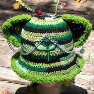 May include: A crocheted bucket hat featuring horizontal stripes in shades of green and black. The hat includes two large, fuzzy green ears, a silver chain draped across the front, and a patch with a green sprout and the text SPROUT ME.