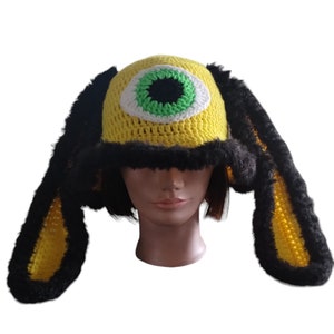 May include: A whimsical, handmade crochet hat in bright yellow, featuring a large, central eye design with green, white, and black details. The hat has long, floppy ears trimmed with black faux fur. The hat is a unique, statement piece.