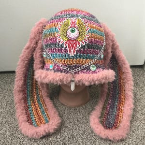 May include: A handmade crochet hat with long, fluffy pink ears. The hat features a colorful striped pattern and is adorned with a silver chain, iridescent gems, and a crystal pendant. A sticker of an eye with wings is on the front.