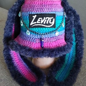 May include: A handmade crochet hat with a bunny ear design. The hat features a vibrant gradient of pink, purple, and blue, with a dark blue faux fur trim. The hat has the word "Levity" on a patch and a decorative chain.