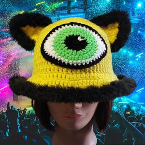 May include: A yellow crocheted hat with a large green eye and black fur trim. The hat is worn by a person with a light complexion and short brown hair.