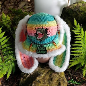 May include: A colorful crochet hat with a rainbow pattern, featuring a dinosaur patch and the text "LOST LANDS". The hat has long, fluffy white ear flaps and is adorned with decorative charms.