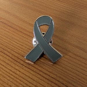Peut inclure: Une épinglette en forme de ruban de sensibilisation gris avec une finition en émail lisse et un contour en métal argenté. Le pin est conçu dans la forme classique en boucle avec deux extrémités inclinées et un fermoir métallique au dos.
