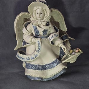 Puede incluir: Figura de ángel color crema con detalles azules, con un sombrero y sosteniendo una cesta de flores. El ángel tiene grandes alas y una pancarta que dice "Grandmothers Like You Are Precious And Few."