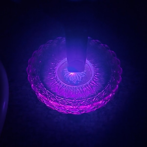 Könnte beinhalten: Ein kreisförmiges Glasobjekt mit strukturiertem Design, das von einem leuchtenden violetten Licht beleuchtet wird. Das Objekt hat einen Stiel und ist vor einem dunklen Hintergrund platziert. Das Glas leuchtet mit einem inneren Licht und erzeugt einen auffälligen visuellen Effekt.