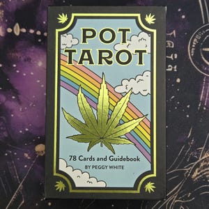 Puede incluir: Caja negra de cartas del tarot con las palabras "POT TAROT" en letras doradas. La caja presenta un arcoíris, nubes y una gran hoja de cannabis verde. La caja también indica "78 cartas y guía" y "BY PEGGY WHITE".