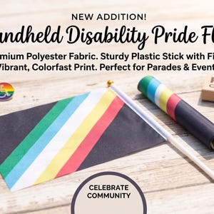 May include: A handheld Disability Pride flag featuring diagonal stripes in red, gold, white, blue, and green on a black background, attached to a white plastic stick with a gold finial. Includes small pins and a rolled flag nearby with text.