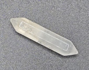 Op de afbeelding: Een heldere kwartskristallen toverstaf met puntige uiteinden. Het kristal is transparant en lijkt gepolijst. De toverstaf ligt op een grijs, getextureerd oppervlak. Geschikt voor kristal genezing en spirituele praktijken.