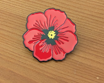 May include: A red enamel pin shaped like a poppy flower with black outlines and a central black circle containing a small yellow stamen. The flower has five distinct petals with subtle embossed lines radiating from the dark centre.