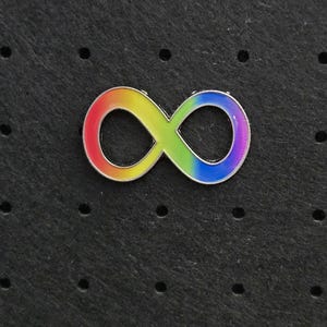 May include: A metal enamel pin shaped like an infinity symbol. The surface of the pin features a smooth gradient of colors transitioning from red to orange, yellow, green, blue, and purple, all outlined with a thin, polished silver-toned metal border.