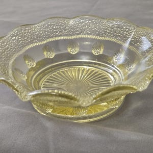 May include: A clear, yellow-tinted glass bowl with a scalloped rim. The rim has a textured, dotted pattern, and the bowl features a sunburst design in the center. Decorative elements include small, shell-like designs.