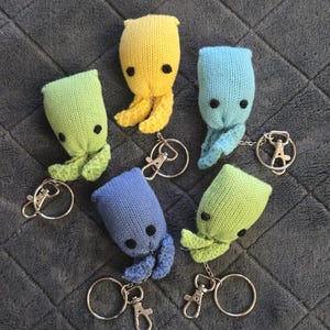 May include: Six knitted octopus keyrings in various colours: yellow, blue, and green. Each octopus has black button eyes and a metal keyring. The keyrings are arranged on a grey, textured surface.