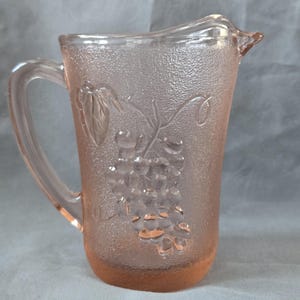 Peut inclure: Pichet en verre rose avec une surface texturée et une poignée. Le pichet présente un motif en relief de raisins et de feuilles. La base du pichet est d'une teinte rose plus foncée. Le pichet est conçu pour servir des boissons.