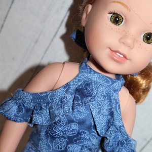 May include: A doll with light skin, freckles, and green eyes wearing a blue denim-look romper featuring a white floral pattern, a halter neckline, and ruffled cold-shoulder sleeves. The garment has a fitted bodice and short bottoms.