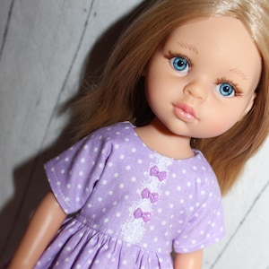 May include: A doll with blonde hair and blue eyes wearing a lavender dress with white polka dots. The dress has short sleeves and is decorated with white lace and purple bows. The doll's face is detailed with painted features.