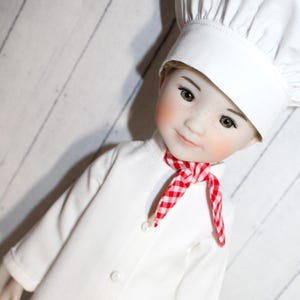May include: A doll dressed as a chef, wearing a white chef's hat, a white button-down shirt, and a red and white checkered neckerchief. The doll has freckles and rosy cheeks, with a slight smile.
