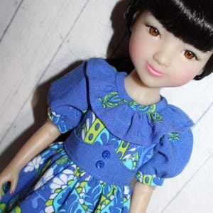 May include: A doll wearing a blue dress with a floral pattern in white, green, and turquoise. The dress has short sleeves and a ruffled collar. The doll has brown eyes and black hair.