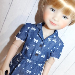 May include: A doll wearing a blue denim dress with white bow patterns. The dress has short sleeves with white lace trim and a button-down front. The doll has blonde hair and blue eyes.