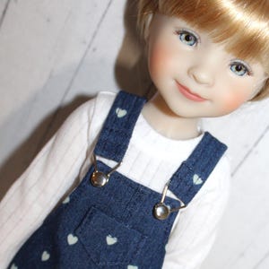 May include: A doll with blonde hair and blue eyes wearing a white long-sleeved ribbed shirt under dark blue denim overalls. The overalls feature a small chest pocket and a repeating pattern of tiny light-colored hearts with silver-toned metal buckles.