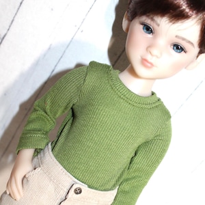 May include: A ball-jointed doll with brown hair and blue eyes, wearing a long-sleeved olive green ribbed knit sweater and beige corduroy pants with a visible button at the waist. The doll has a fair complexion and a neutral facial expression.
