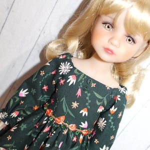 May include: A doll wearing a dark green dress with a floral pattern. The dress features white, pink, and orange flowers, with a decorative trim around the waist. The doll has blonde hair and is looking at the viewer.