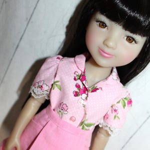 May include: A doll with brown eyes and black hair wearing a pink outfit. The outfit includes a pink blouse with a floral pattern and a matching pink skirt. The blouse has white lace trim on the sleeves.