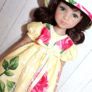 May include: A doll wearing a yellow dress with white polka dots and a floral pattern of pink roses. The dress has a bow at the waist and a white lace trim. The doll has brown hair, green eyes, and a matching hat with a floral design.