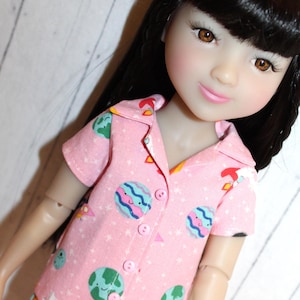 May include: A pink pajama top with a short sleeve and a button-up front. The pajama top has a white background with a repeating pattern of planets, stars, and rockets.