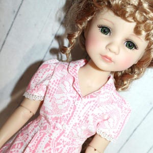 May include: A doll with green eyes and curly blonde hair wearing a pink and white patterned dress. The dress has a collared neckline and short sleeves with lace trim. The doll's face is detailed with painted features.
