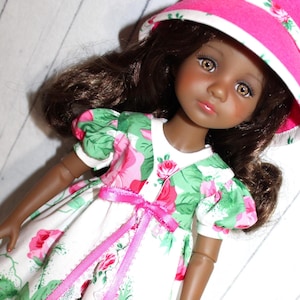 May include: A doll with brown hair and brown eyes wearing a white dress with a pink floral pattern and green leaves. The dress has a pink ribbon bow. The doll is wearing a pink hat with a white floral trim.
