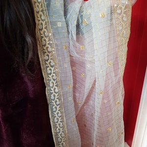 Puede incluir: Un dupatta blanco de gasa transparente con un borde bordado en oro y plata. El dupatta tiene un patr&oacute;n de cuadr&iacute;cula y est&aacute; decorado con peque&ntilde;as lentejuelas doradas.